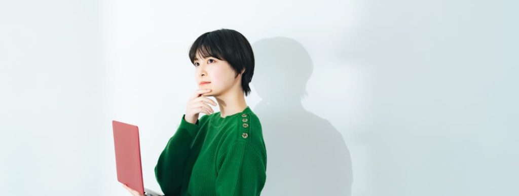 赤いノートパソコンを持ち、悩ましい表情を浮かべるフリーランスエンジニアの女性。