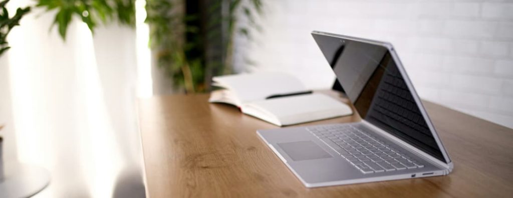 木製のデスクに置かれたノートパソコンと、開いたノート、ペン。背景には観葉植物がある。