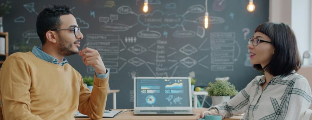 図解が描かれた黒板とデータが表示されたPCを前に、戦略を練る男女のビジネスパーソン。