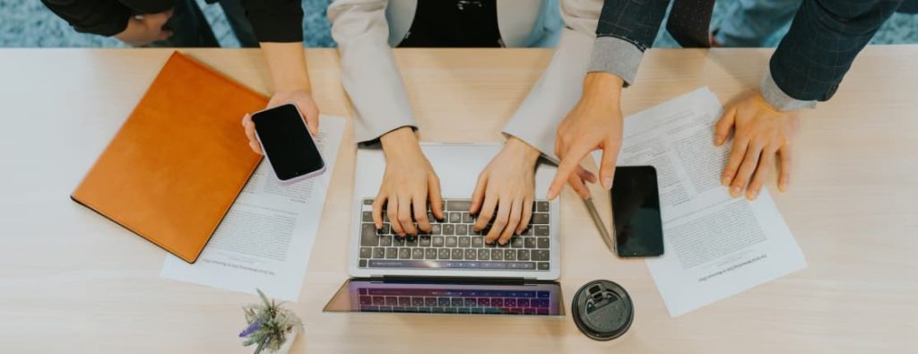 デスク上のノートPCやスマートフォン、書類を指さしながら確認を行う人々。