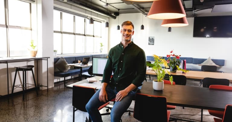 開放的なコワーキングスペースで、デスクの端に腰掛けて微笑む、緑のシャツを着た男性。