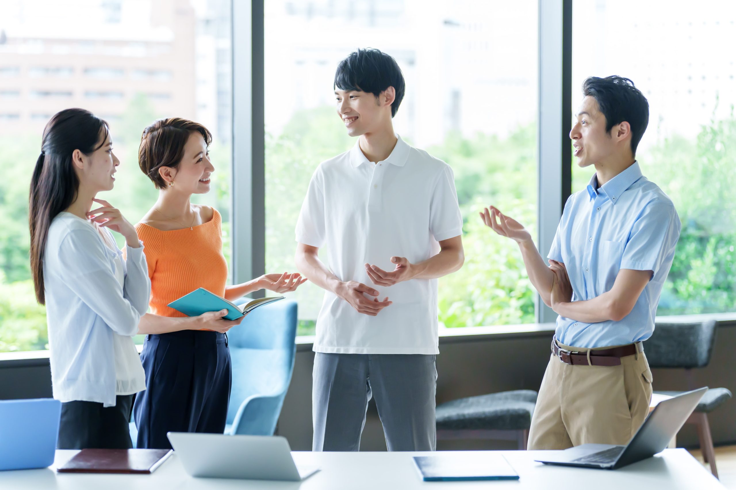 カジュアルな服装の男女4人がオフィスで談笑する様子。机にはノートパソコンが置かれている。