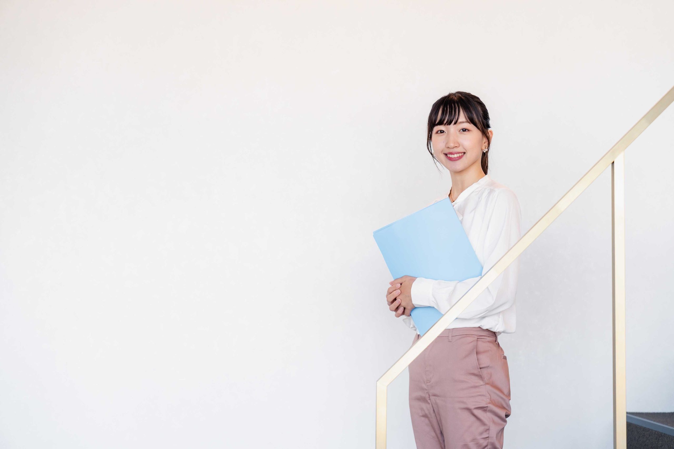 笑顔で階段に立つ若い女性の全身写真。白いブラウスとピンクのパンツを着用し、水色のファイルを抱えている。