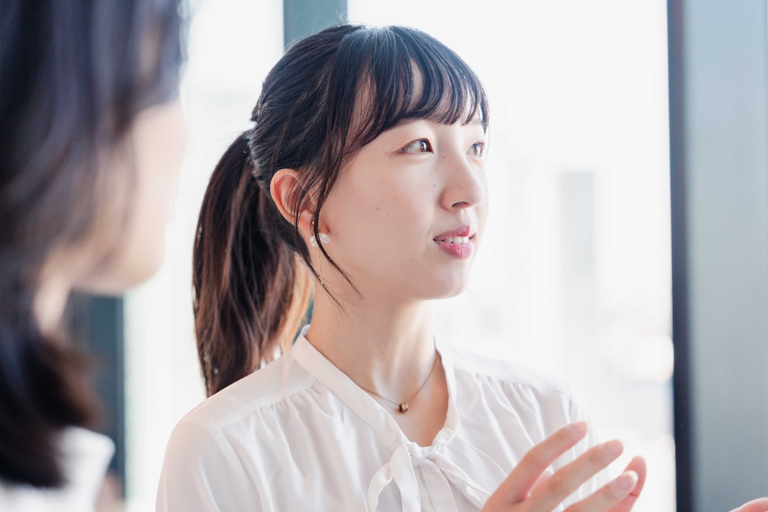 白いブラウスを着たポニーテールの若い女性が、誰かに向かって身振り手振りを交えながら熱心に話している様子。